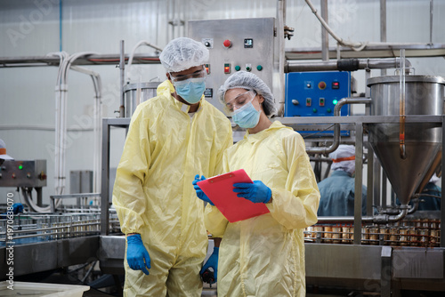 Two industrial engineer supervisors inspect and examine quality control, production process at automate conveyor in canned food manufacturing factory, and protective uniforms for hygiene operations.
