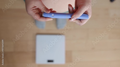 A horizontal close up of a young woman screwing a needle on pre-filled pen injector with semaglutide to prepare it for taking medicine on floor and scales background
