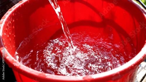 A stream of clear water is being poured into a bright red plastic container, filling it up