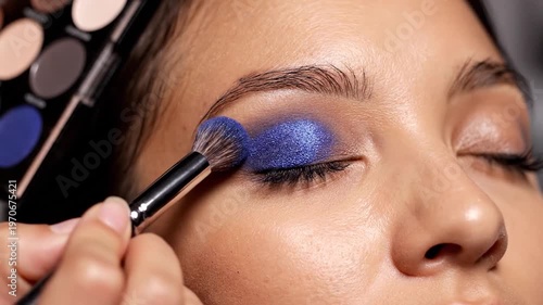 A makeup artist applies vibrant blue eyeshadow to a person's eyelid using a brush from a palette of colors