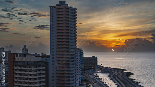 Sunset in a seaside town. The sun is low over the ocean, shining behind the clouds. The evening sky is highlighted in gold near the horizon. The highway along the embankment. Multi-storey buildings 

