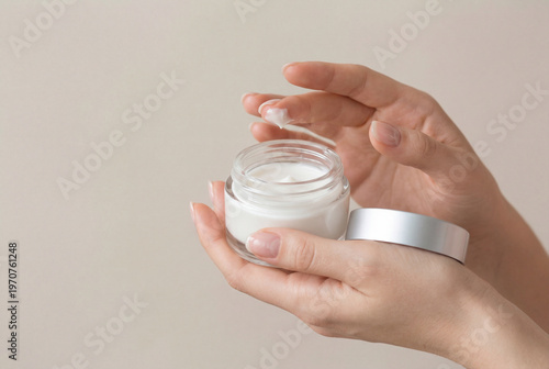 Woman hands holding open glass jar of white moisturizing cream against beige background