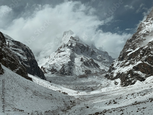 Fotografi A massive snow-covered Himalayan mountain peak towering over a rugged glacial valley under a dark dramatic sky