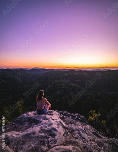 Woman sits on a rock, enjoying a vibrant sunset over a vast forest landscape.