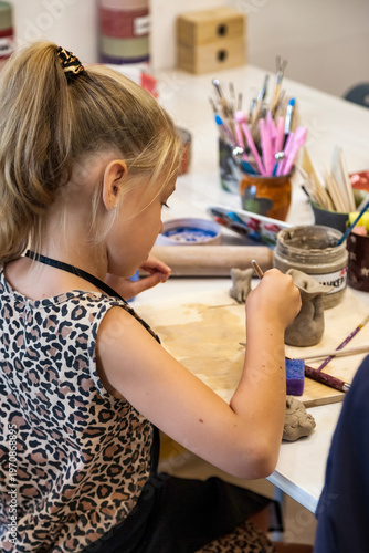 A young girl is diligently working with clay at a white table. She is shaping it with tools in a bright and creative workshop indoors. She is enjoying her time in an art class.