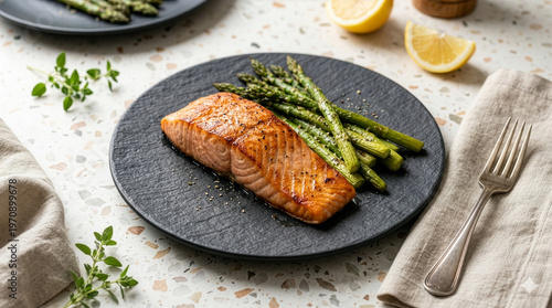 Premium roasted salmon fillet with visible grill marks and moist texture served with roasted green asparagus spears on a dark slate plate, placed on a terrazzo table with folded linen napkin and lemon
