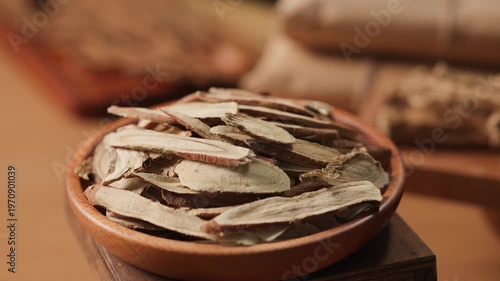Thin slices of licorice root arranged in a wooden bowl. Light golden tones and smooth cut surfaces are emphasized through rotation, highlighting the dried herbal texture.