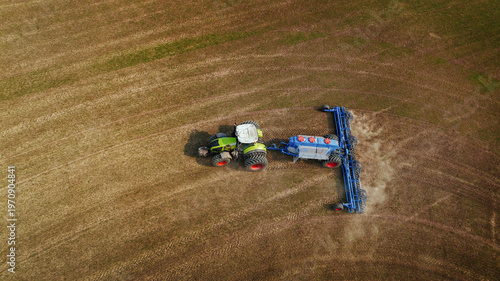 Aerial view of green tractor with blue plow working on brown agricultural field, creating furrows in soil during farming season