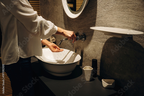 Woman washing hands at modern bathroom sink with round mirror, stylish decor, and minimalist design elements in a contemporary setting