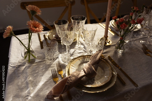 Elegant dinner table setting with gold cutlery, crystal glasses, and pink flowers on a white linen cloth. Warm sunset light creates a romantic atmosphere for a wedding, banquet, or festive celebration