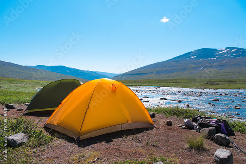 Bright orange camping tent pitched by a crystal clear mountain stream in the vast Sweden nature landscape while trekking the Kungsleden trail in Arctic Lapland