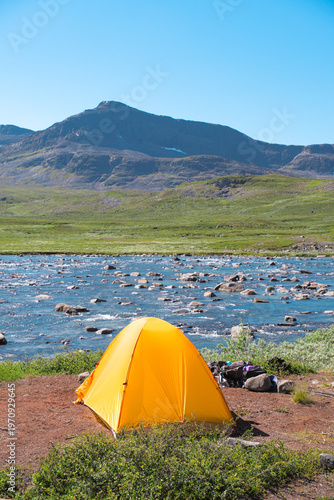 Bright orange camping tent pitched by a crystal clear mountain stream in the vast Sweden nature landscape while trekking the Kungsleden trail in Arctic Lapland