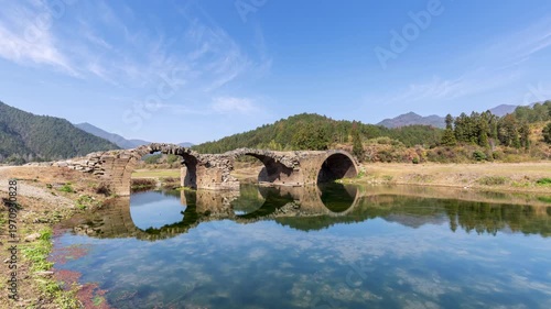 Time-lapse of the weathered three-arch stone bridge under blue sky, this ancient structure exemplifies traditional architectural beauty and cultural heritage. Wuyuan County, Jiangxi Province, China