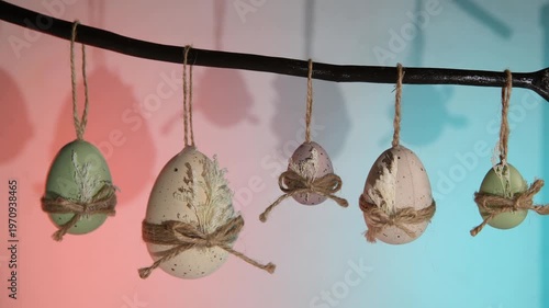 Pastel easter eggs in green, white, and purple hang from a natural branch, tied with twine bows and dried leaves against a vibrant background for spring celebration