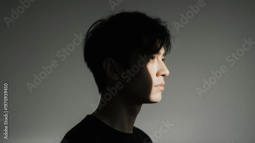 Cinematic profile portrait of a young Asian man with tousled dark hair against a gray background. Low-key lighting creates a dramatic contrast with light hitting only the face.