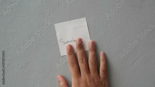 Hand Placing a White Sticky Note with Subscribe Written on it to a Textured Gray Wall
