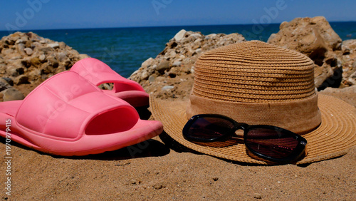 Straw hat and pink rubber slippers on a sandy beach with blue sea in the background. Concept of summer vacation, seaside relaxation, travel, and family holiday. Sunny coastal lifestyle and leisure.