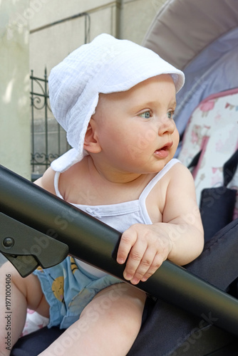 A child enjoys the outdoors while sitting in a stroller. The child wears a white hat and looks around. It is a sunny day in a city setting.