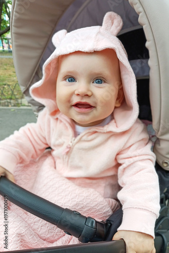 A young child with blue eyes smiles while sitting in a stroller. The kid wears a pink outfit and is outside in a park on a sunny day.
