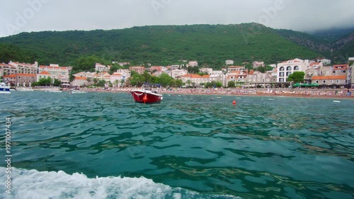 view from a tourist boat sailing into the sea to the resort town against the backdrop of mountains covered with forest, Petrovac, Montenegro, city beach and hotels, beautiful coastline
