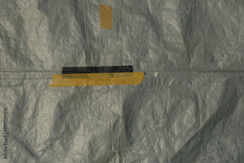 High angle view of weathered grey plastic tarpaulin with adhesive tape repairs. Close up of a wrinkled grey waterproof tarp texture featuring yellow and black duct tape patches. Industrial background 