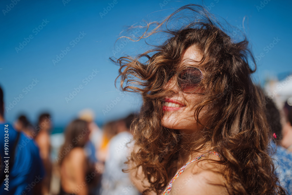 Naklejka premium Close-up of joyful woman with windblown hair at oceanfront party, people dancing blurred in background, bright blue sky, high detail, crisp natural lighting, advertising-ready composition, balanced
