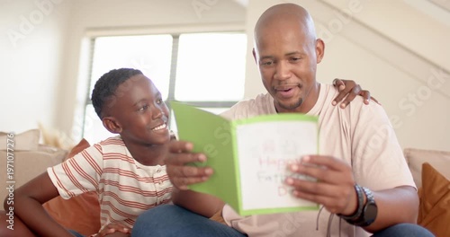 African dad, teen son after giving green handmade card on couch, dad reading, hugging, showing love