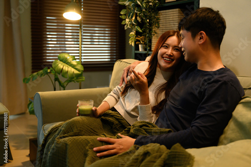 Bild auf Leinwand Asian couple relaxing on a sofa, watching TV together with smiles, enjoying a cozy and intimate evening at home