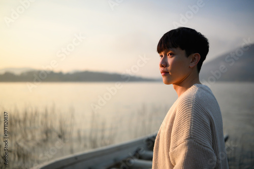 Fotografi Close portrait of a young Asian man standing by a peaceful lake at sunrise, look