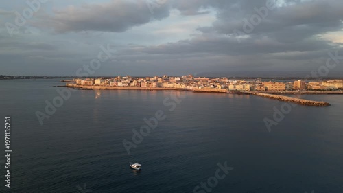 Fishing boat navigates the calm waters off the coast of Sicily, with a picturesque coastal town and rocky shoreline illuminated by the warm evening light