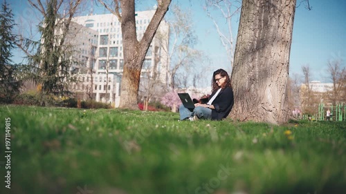 Young student woman in glasses working on a laptop under a tree in a blooming spring park. Outdoor studying, education, focus, and modern student lifestyle concept.