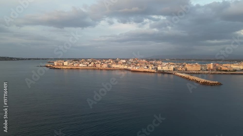 Sunrise panorama of Ortigia, Sicily, showcasing the tranquil waters and historic architecture along the coastline with soft clouds in the sky reflecting morning light