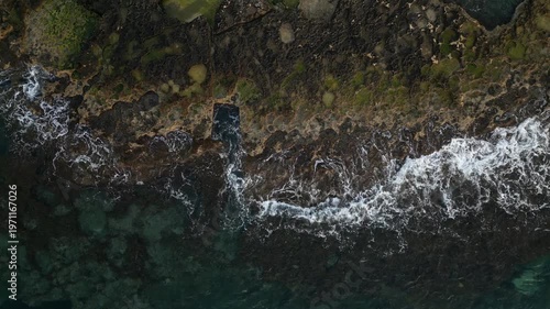 Aerial view of rocky coastline at sunrise with crashing waves, revealing vibrant ocean colors and textures along the shore, showcasing natural beauty and serenity