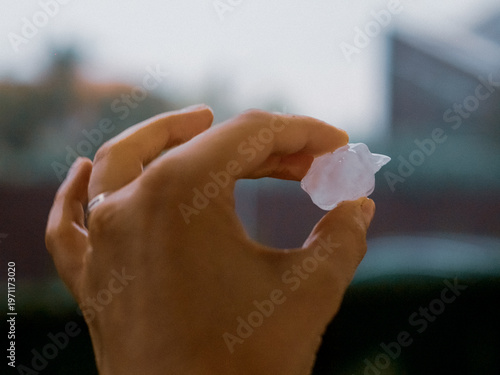 A woman's hand displays a large ice cube right after a hailstorm in the neighborhood. The sky is cloudy and the ground is wet from the storm.