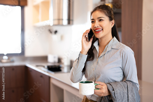  Young business woman holding suits having a coffee and business talking on smartphone in kitchen while getting ready to go to work in the morning.