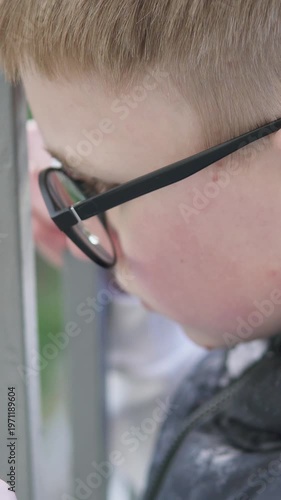 A teenage boy stands behind a metal fence, looking upset and isolated. The scene conveys sadness and bullying at school, highlighting emotional struggle and vulnerability. Vertical video.