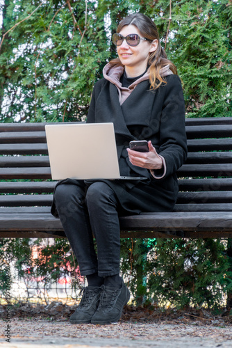 Woman working remotely with laptop and phone smiling on bench digital nomad