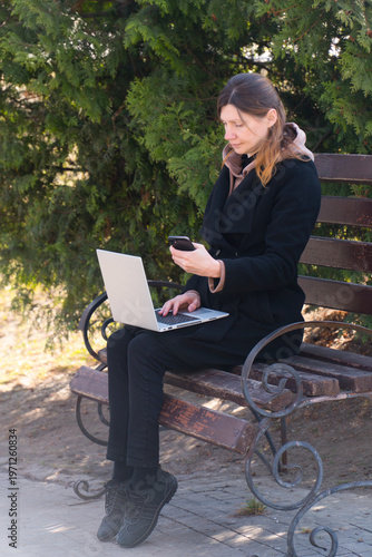 Woman using laptop and smartphone for remote freelance business on park bench