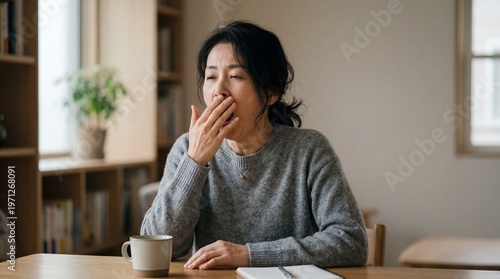 A tired Asian woman sits at a table in her home, yawning with her hand covering her mouth, next to a mug and notebook.