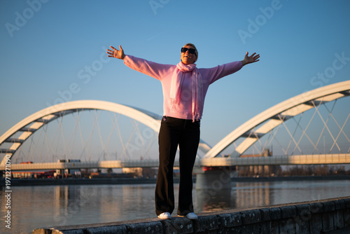 Carefree woman standing on quayside with arms outstretched, feeling inspired and relaxed, embracing city life, sunlight and peaceful river atmosphere.