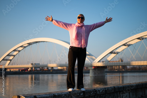 Carefree woman standing on quayside with arms outstretched, feeling inspired and relaxed, embracing city life, sunlight and peaceful river atmosphere.