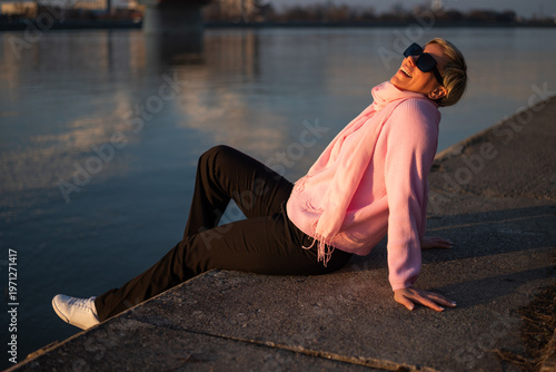 Happy urban woman with sunglasses smiling while sitting and relaxing by the city river on a beautiful sunny day.