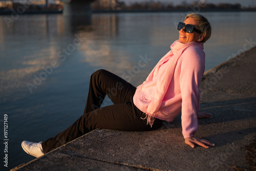 Portrait of happy and carefree urban woman with short blonde hair sitting by the city river on a sunny day.