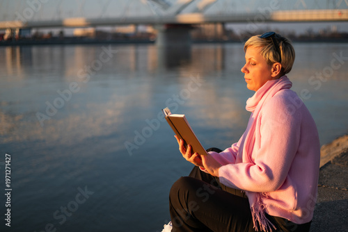 Blonde woman with a short hair reading a book while enjoying sitting comfortably by river in the city. Serene, contemplative mood with bridge and city life in the background, bathed in warm sunlight.