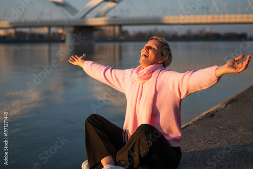 Beautiful and modern blonde woman with a short hair sitting by river with arms raised and eyes closed, enjoying sunlight and freedom.