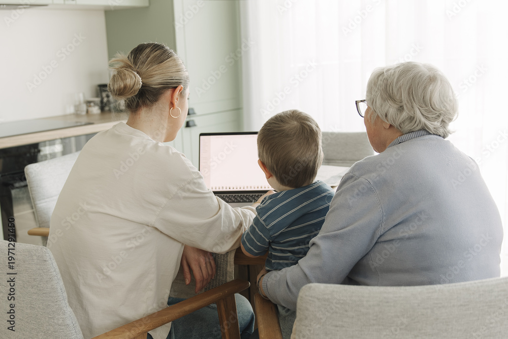 Obraz premium Three generations sitting together at home using laptop. Young caregiver helping senior woman and child with digital technology. Concept of family support, intergenerational learning and modern lifest