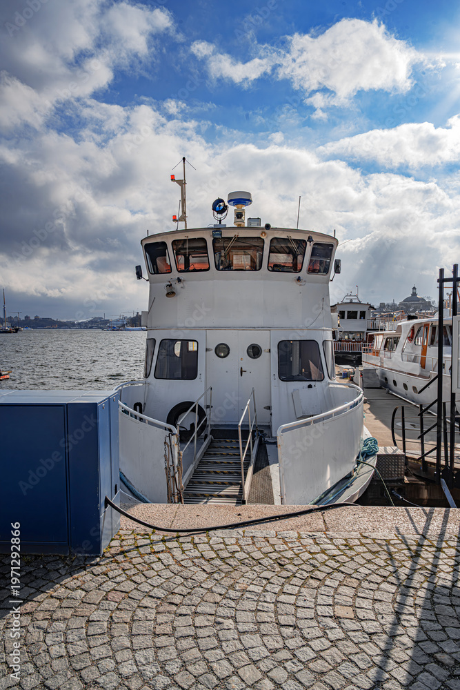 Fototapeta premium A white passenger ferry sits docked at a cobblestone pier under a bright, cloudy sky in Stockholm harbor.