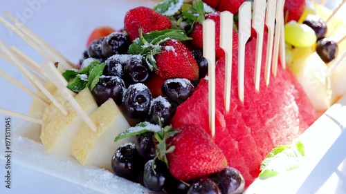 Colorful fruit platter featuring watermelon, strawberries, cherries, pineapple, grapes, and melon, garnished with mint and powdered sugar, presented with wooden skewers