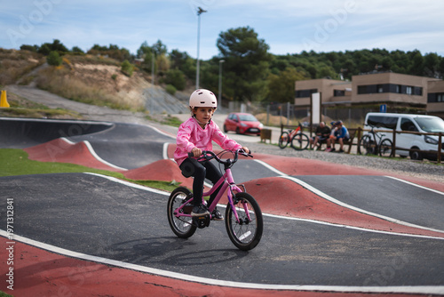 A young girl riding a pink bicycle on a paved bike track with a helmet on her head