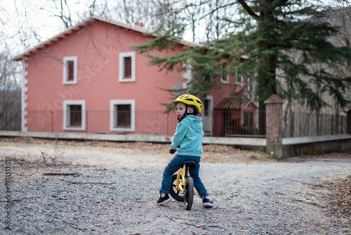 A young child riding a bike in front of a pink house with a yellow helmet on a gravel road